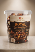 A clear plastic tub of TRS The Royal Snacks Royal Choco Caramel Cured Gourmet Popcorn sits on a light beige countertop under a warm spotlight against a beige wall.